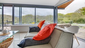 A modern living room with a sofa, a glass coffee table, and a view of a garden through large windows. - 4 Bed in Barmouth (oc-bow259) (Barmouth)