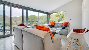 A modern living room with large windows, a sofa, a chair, and a glass coffee table. - 4 Bed in Barmouth (oc-bow259) (Barmouth)