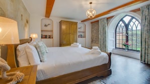 Une chambre à coucher avec un grand lit, des poutres en bois, un lustre, une bibliothèque et une fenêtre donnant sur un beau paysage.
