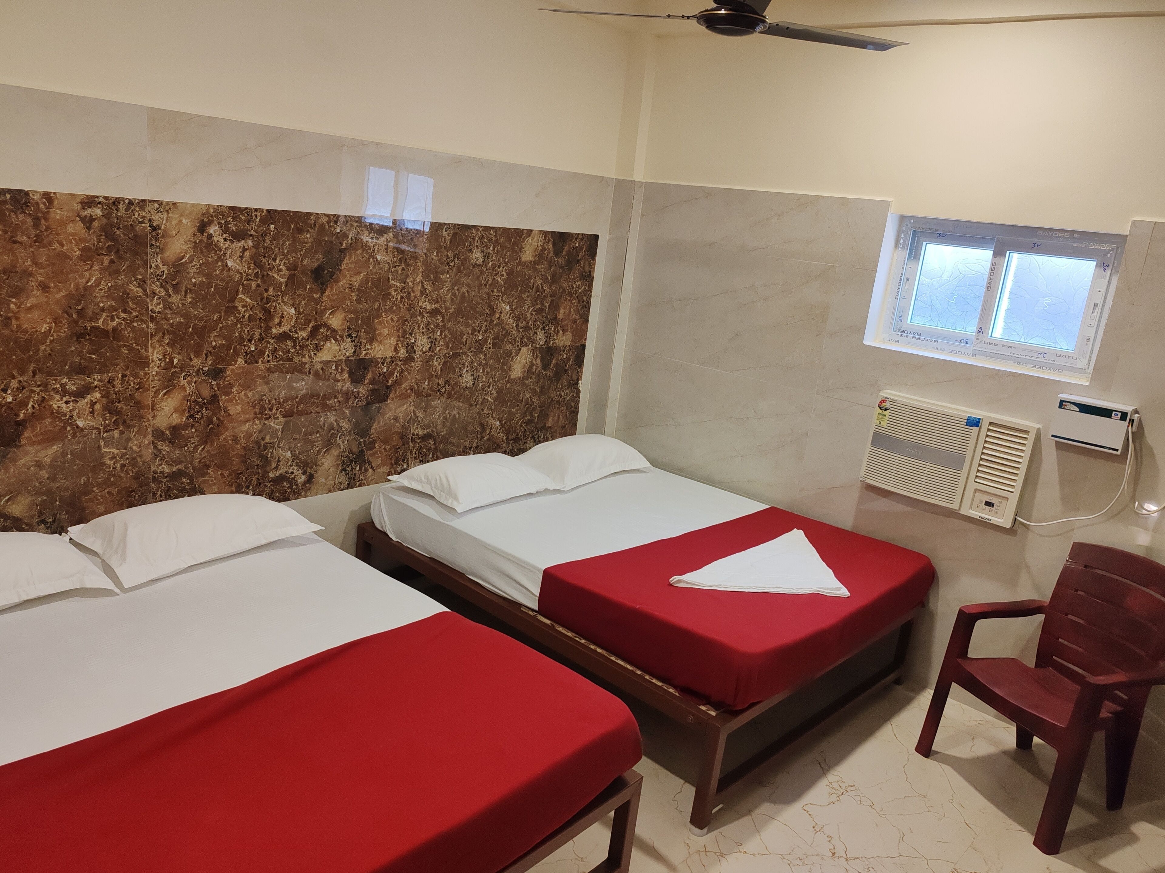 Habitación de hotel con dos camas, una silla roja, ventilador de techo y ventana con vistas.