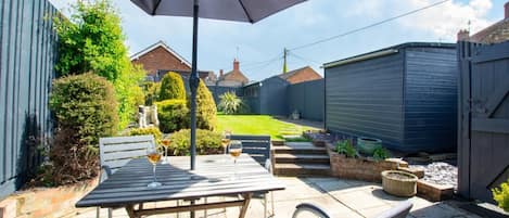 An outdoor patio with a table and chairs, a large umbrella, and a small garden area with a lawn and shrubs.
