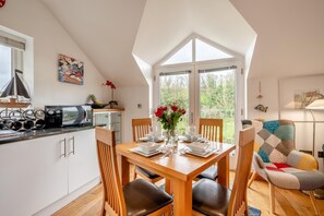 A dining area with a wooden table set for four, surrounded by chairs, a kitchen area with a microwave and cabinets, and a large window offering a view of greenery. - 2 Bed in Wadebridge (oc-p00445) (Little Petherick)