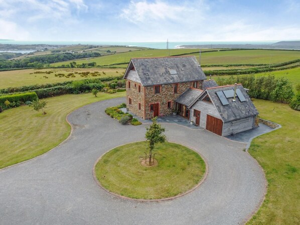 A stone house with a sloped roof and a circular driveway, surrounded by green fields and a view of the sea. - 4 Bed in Instow (oc-h32283) (Westleigh)
