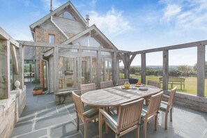 A covered patio with a wooden table and chairs, a grill, and a view of a house in the background. - 4 Bed in Instow (oc-h32283) (Westleigh)