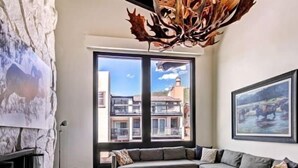 Een moderne woonkamer met een grijze bank, een houten salontafel en een groot raam met uitzicht op gebouwen en een heldere lucht.