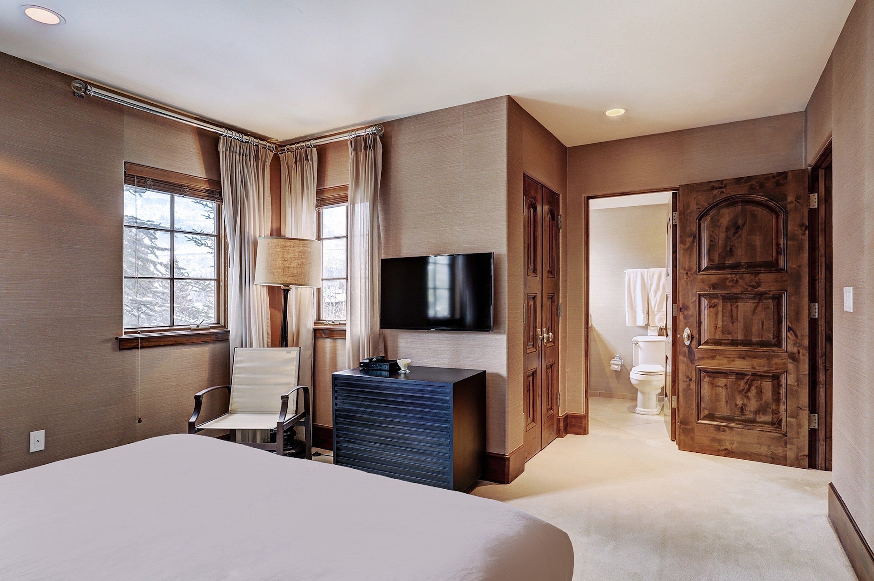 A hotel room with a large bed, a desk with a lamp, a TV, and a wardrobe. There is a window with curtains and a view of trees outside.