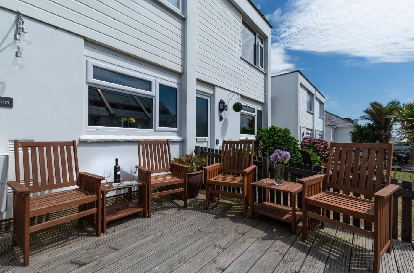 A wooden deck with outdoor furniture, including chairs and a table, in front of a white building with windows and a door.