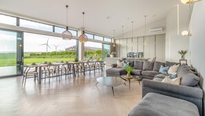 A spacious living room with a large dining area, modern furniture, and large windows offering a view of wind turbines. - 7 Bed in Marwood (oc-w32673) (Marwood)