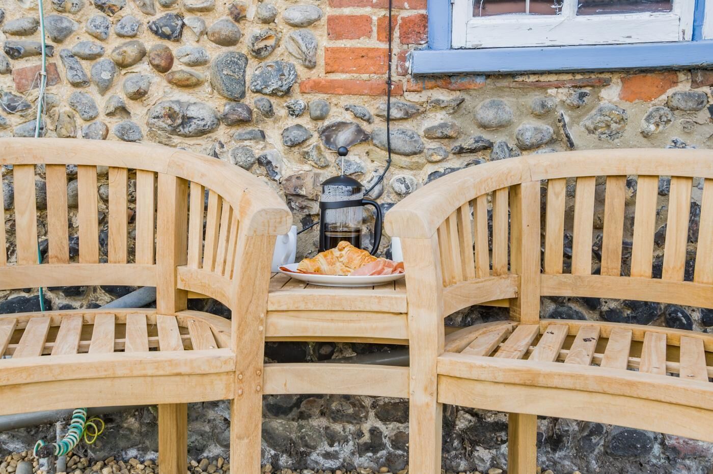 Two wooden chairs with slatted backs and a table in front, a plate with croissants, and a coffee maker on the table.