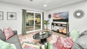 A living room with a ceiling fan, a glass door leading to a balcony, a large painting, and a round wall clock. - 4 BR Luxury Home in Friendswood (Friendswood)