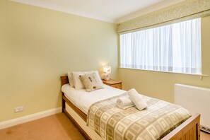 Ein Schlafzimmer mit Bett, Nachttisch mit Lampe, Fenster mit Vorhängen und Heizkörper.