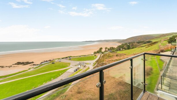 Udsigt over kysten fra en balkon med strand, grønne områder og en mindre bygning.