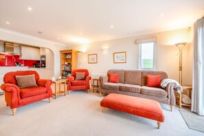 Ein Wohnzimmer mit einem roten Sessel, einem braunen Sofa und passendem Hocker. Ein Bücherregal und ein kleiner Tisch mit Büchern sind ebenfalls zu sehen.