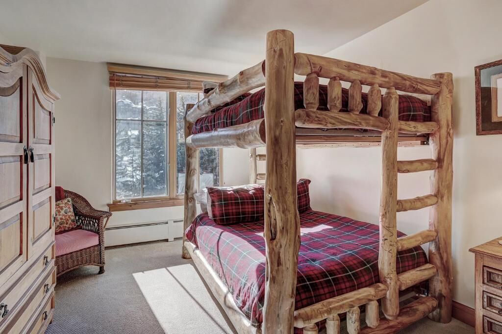 A rustic wooden four-poster bed with plaid bedding, a wicker chair, and a wooden dresser in a cozy room with a large window.