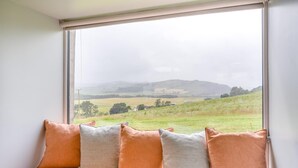 A room with a large window overlooking a grassy field and hills. There is a bed with a beige mattress and four pillows in shades of orange and gray. - 1 Bed in Newburgh (oc-h32593) (Newburgh)