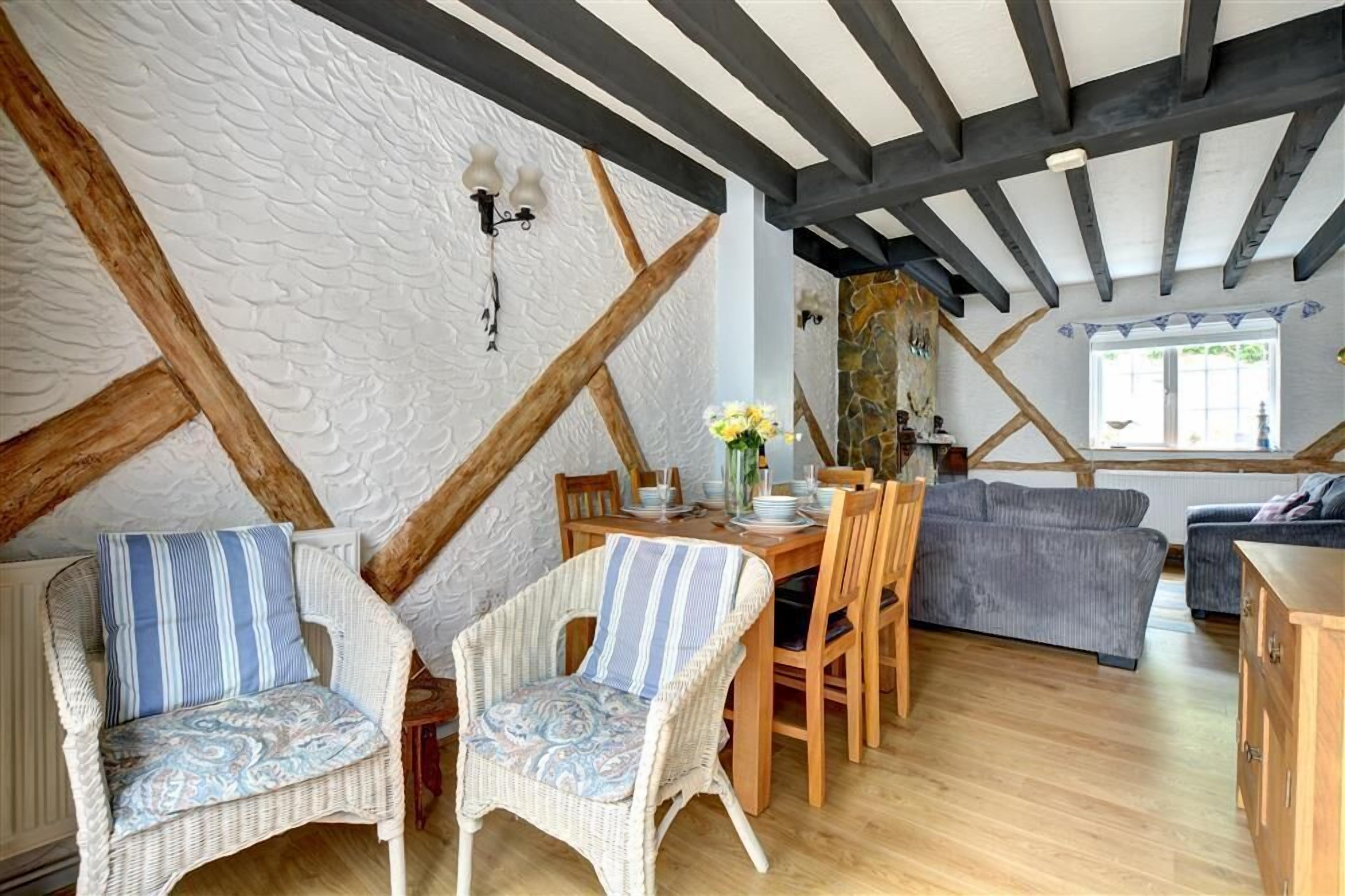 Une pièce chaleureuse avec des poutres apparentes, un mur en pierre et un coin repas agrémenté de chaises en osier et d’une table dressée pour deux personnes.
