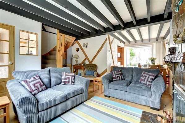 A cozy living room with two grey striped sofas, a wooden staircase, and a rustic kitchen area. - 3 Bed in Newquay (oc-p00484) (Newquay)