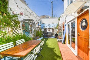 A narrow courtyard with a wooden table and benches, a small lawn, and a door with a ship's wheel design. - 3 Bed in Newquay (oc-p00484) (Newquay)