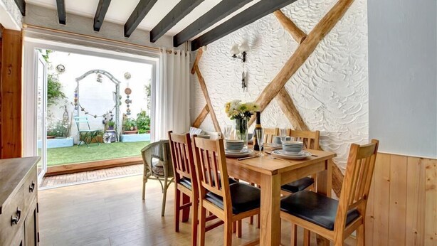 Een eetgedeelte met een houten tafel voor vier personen, omringd door houten stoelen met zwarte kussens. Een witte, gestructureerde muur en een houten balk in het plafond zijn zichtbaar.
