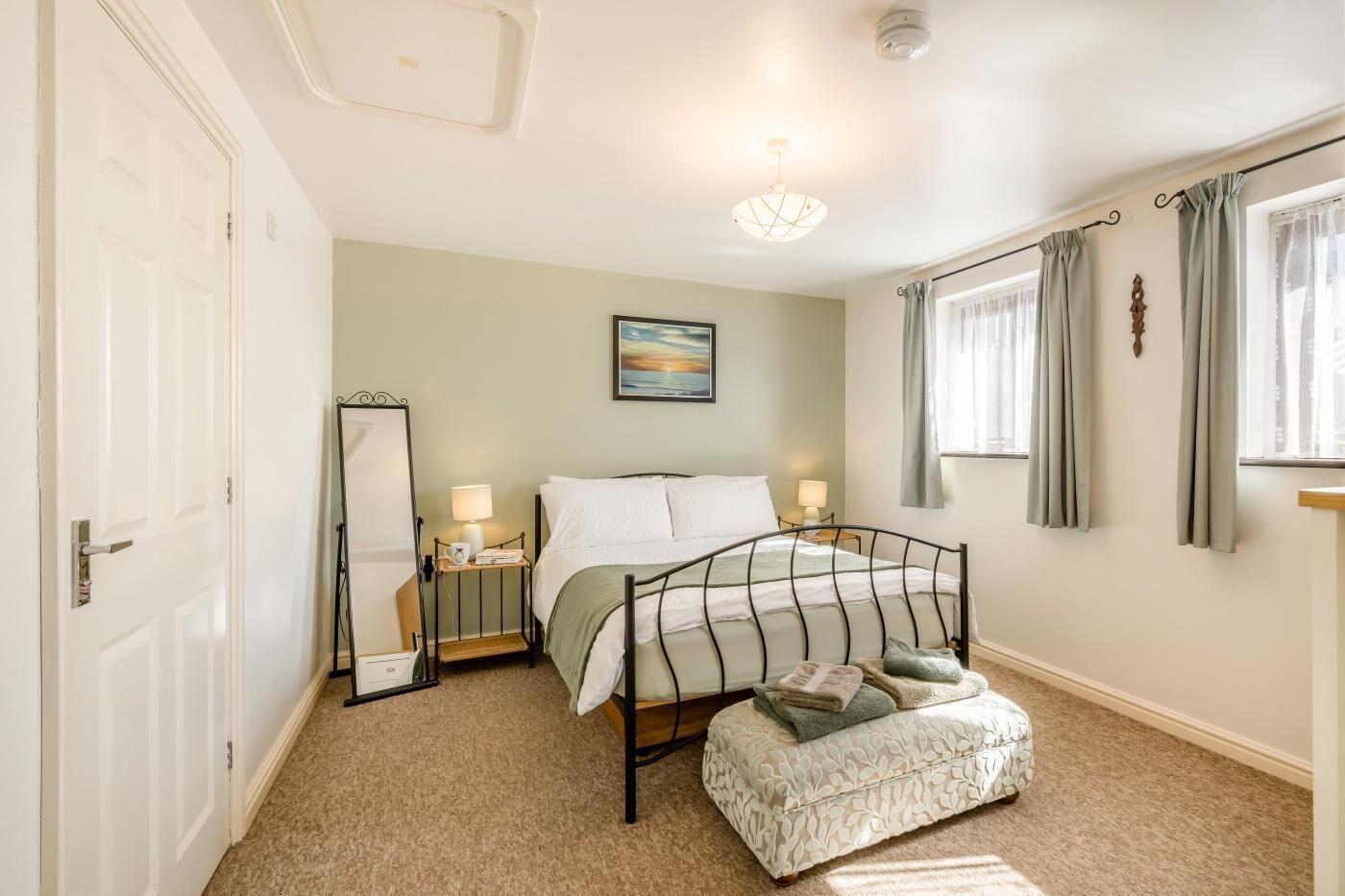 A bedroom with a bed, bedside tables, a mirror, a window with curtains, and a ceiling light fixture.