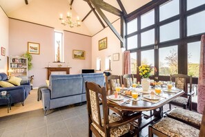 A dining area with a table set for breakfast, a blue sofa, and a bookshelf in a room with large windows. - 3 Bed in Little Ellingham (oc-2147) (Little Ellingham)