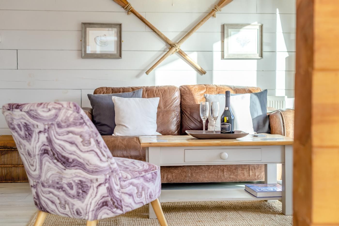 A cozy living room with a brown leather sofa, a purple marble chair, and a wooden coffee table with two glasses and a bottle.