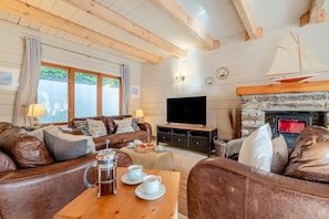 A cozy living room with a leather sofa, a coffee table with a coffee pot and cups, a flat-screen TV, and a stone fireplace. - 3 Bed in Wadebridge (oc-p00104) (Little Petherick)