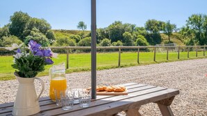 A picnic setup with a table, a pitcher of orange juice, a vase of flowers, and a croissant on a wooden bench. - 4 Bed in Welshpool (oc-wab267) (Welshpool)