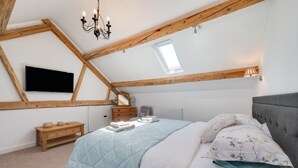 A bedroom with a sloped ceiling, wooden beams, a bed with a quilt, a bedside table, a dresser, a mirror, a TV, and a window. - 4 Bed in Welshpool (oc-wab267) (Welshpool)