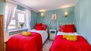 A bedroom with two beds, each with red bedding and white headboards. There are two bedside tables with lamps, a window with white curtains, and a vase with flowers on the windowsill. - 2 Bed in Cromer (oc-t32572) (Cromer)