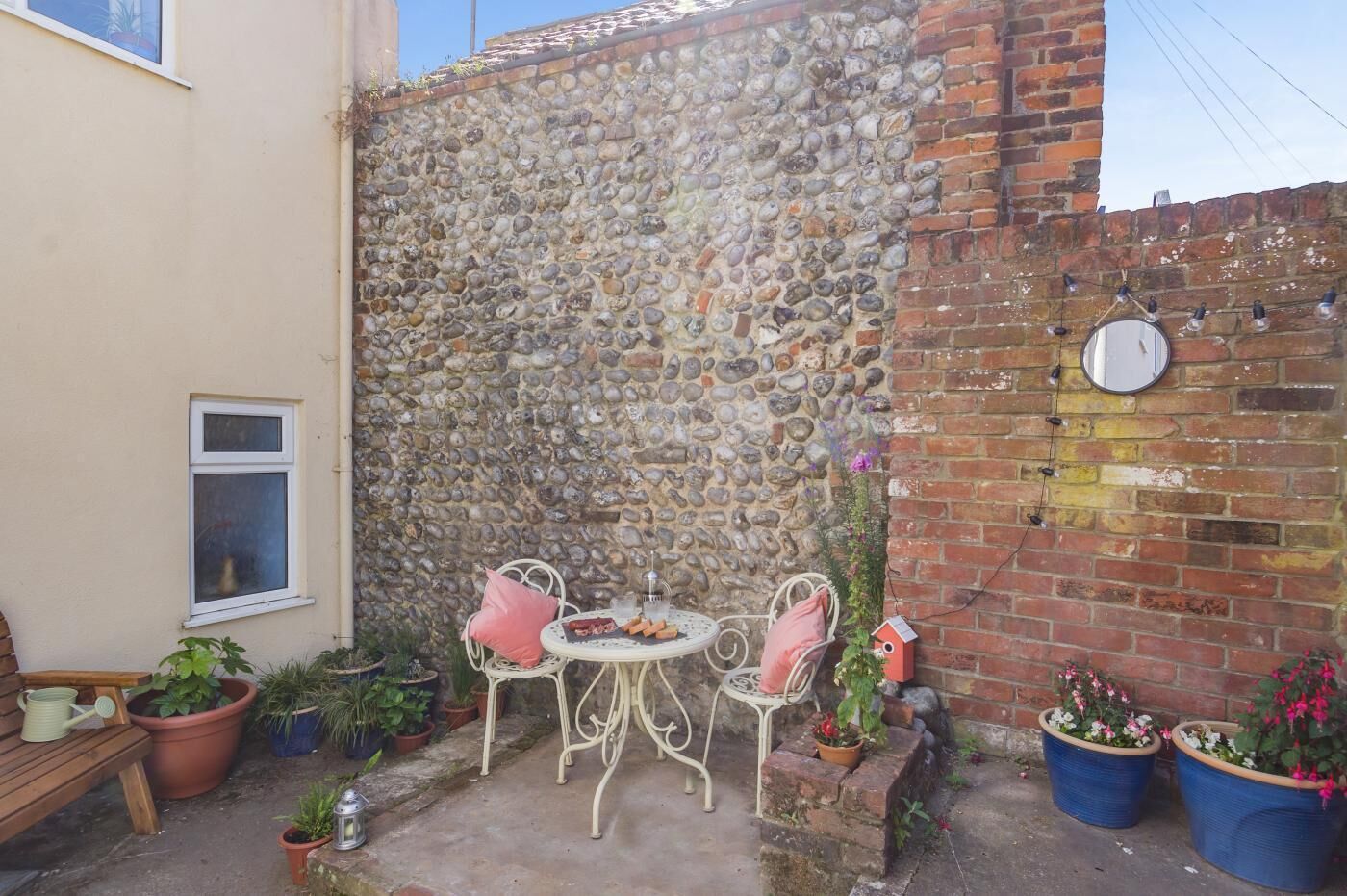 Un mur de pierre, un mur de briques, une fenêtre, une table avec des chaises et des plantes en pot.