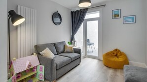 Un saló modern amb un sofà gris, un seient de gerro amb color groc, una taula petita amb una làmpada, un rellotge de paret i una finestra amb cortines.