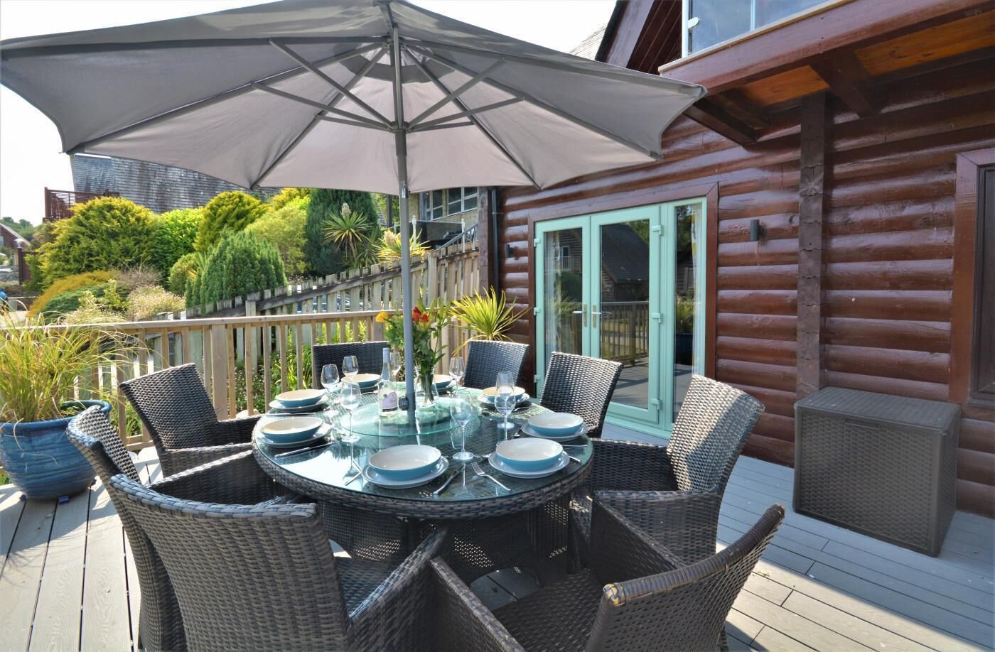 A wooden deck with a glass table set for dining, surrounded by wicker chairs, and a large umbrella providing shade.