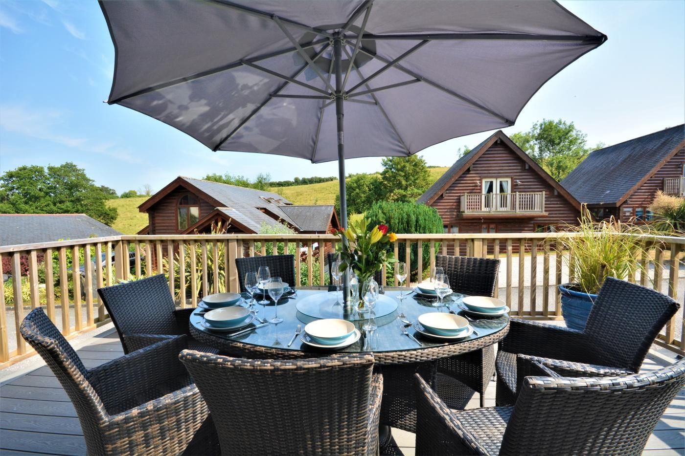 A deck with a table set for dining, surrounded by wicker chairs, under a large umbrella.