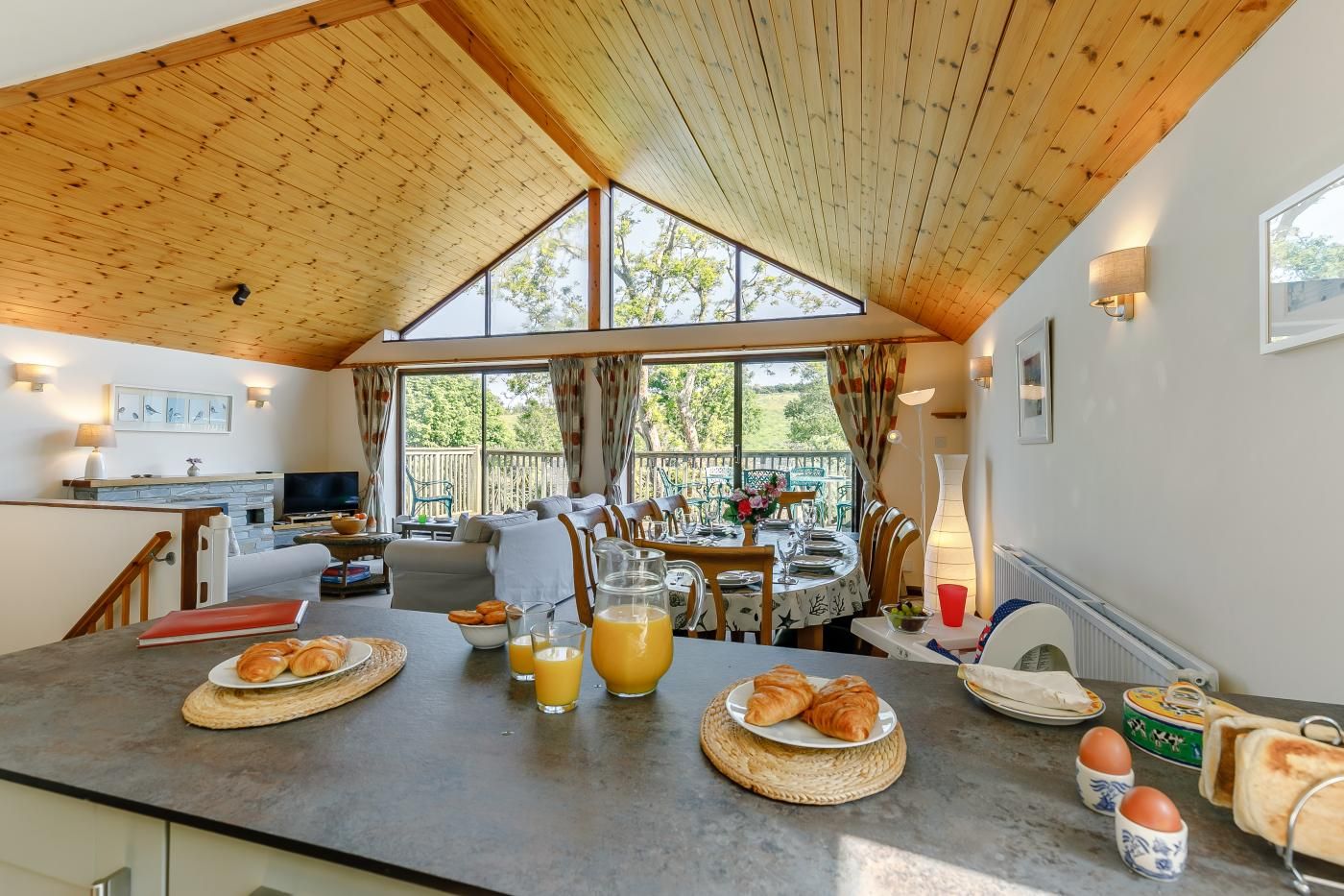 Un desayuno acogedor con cruasanes, zumo de naranja y huevos sobre una mesa de madera en una habitación luminosa con un ventanal y vistas a la vegetación.