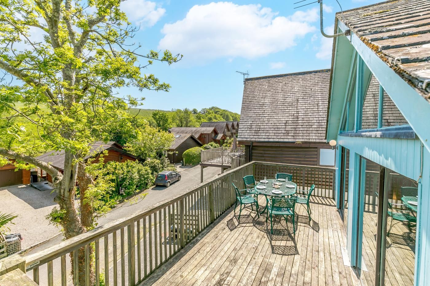 A wooden deck with a table and chairs, surrounded by a wooden fence, overlooking a residential area with houses and a car parked on the street.