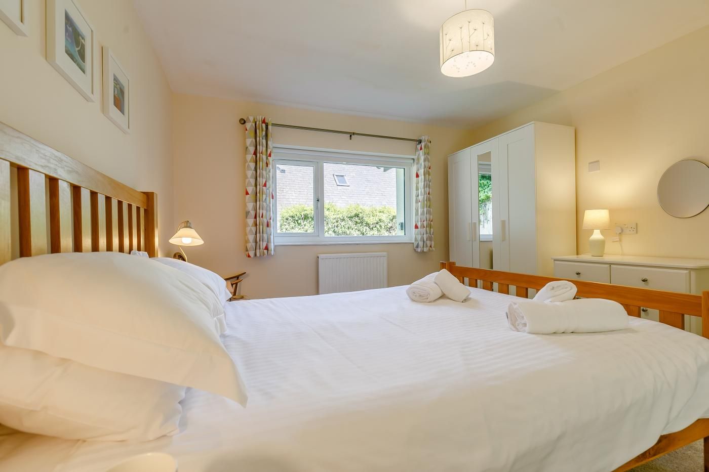 A neatly made bed with pillows and towels, a bedside lamp, a window with curtains, a radiator, a wooden headboard, a mirror, and a white dresser.