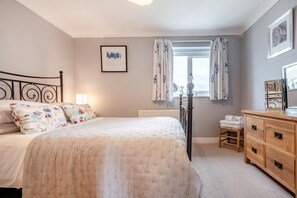 Ein Schlafzimmer mit Bett, Nachttischen, einem Schminktisch, einem Fenster mit Vorhängen und einem gerahmten Bild an der Wand.
