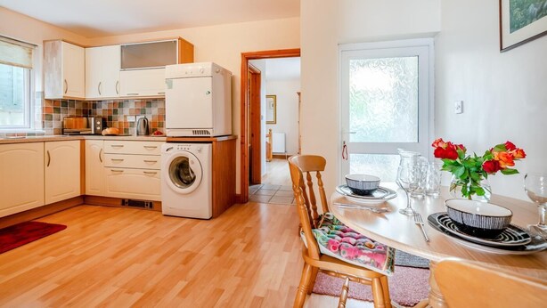 A compact kitchen with a washing machine, a dining area with a table set for two, and a view of the outdoors. - 1 Bed in Wadebridge (oc-p00232) (Wadebridge)