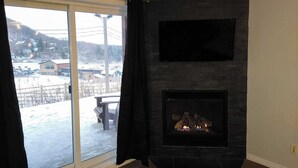 Una habitación con chimenea, televisor montado encima y un amplio ventanal con vistas a un paisaje nevado.
