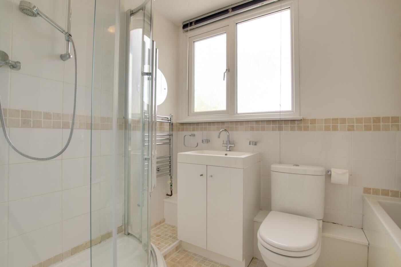 Een badkamer met een douche met glazen wand, een witte wastafel, een toilet en een raam boven de wastafel.