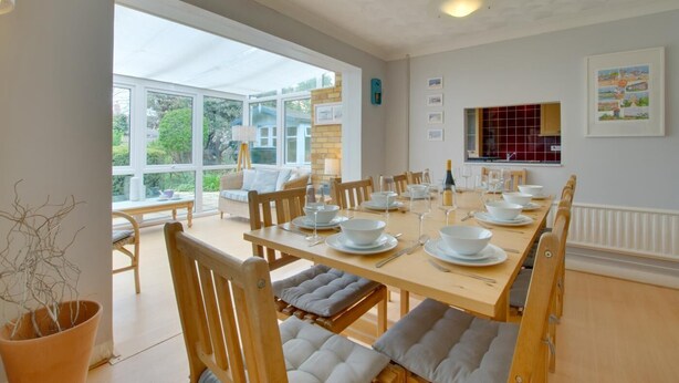 A dining area with a long table set for six, surrounded by wooden chairs with cushioned seats. - 4 Bed in Reydon (oc-fll) (Reydon)