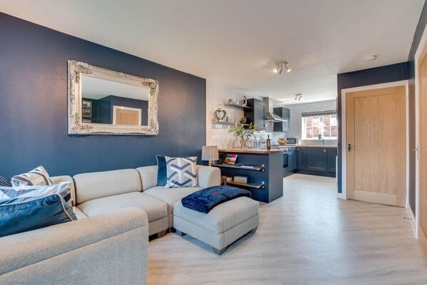 A modern living room with a sofa, ottoman, and a coffee table. There is a kitchen area with a countertop and cabinets. A wooden door is visible in the background. - 2 Bed in Whitby (oc-w1008) (Whitby)