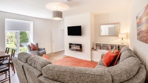 A modern living room with a grey sofa, a flat-screen TV, a fireplace, and a large window overlooking a garden. - 3 Bed in Morpeth (oc-t32249) (Thirston)