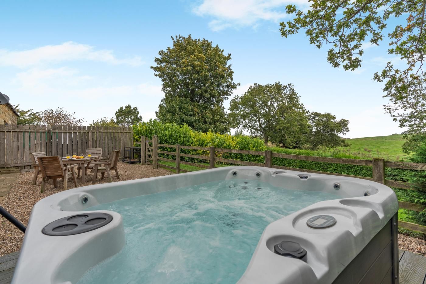 A hot tub with a wooden deck and chairs, surrounded by a wooden fence and greenery.