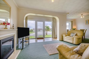 A living room with a fireplace, a television, a sofa, and a large sliding glass door leading to a patio. - 3 Bed in Padstow (oc-p00131) (Saint Merryn)