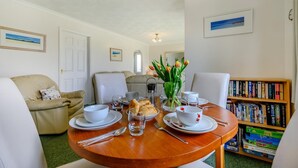 Een eethoek met een ronde houten tafel gedekt voor twee personen, omringd door witte stoelen. Borden, kommen, kopjes en een vaas met tulpen staan er. Achtergrond toont een boekenkast met boeken.