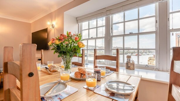 Ein Essbereich mit einem für das Frühstück gedeckten Tisch, einer Blumenvase und Blick auf den Hafen durch große Fenster.