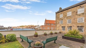 Eine Uferterrasse mit zwei grünen Bänken, Blumentöpfen und Blick auf den Hafen sowie Gebäude im Hintergrund.