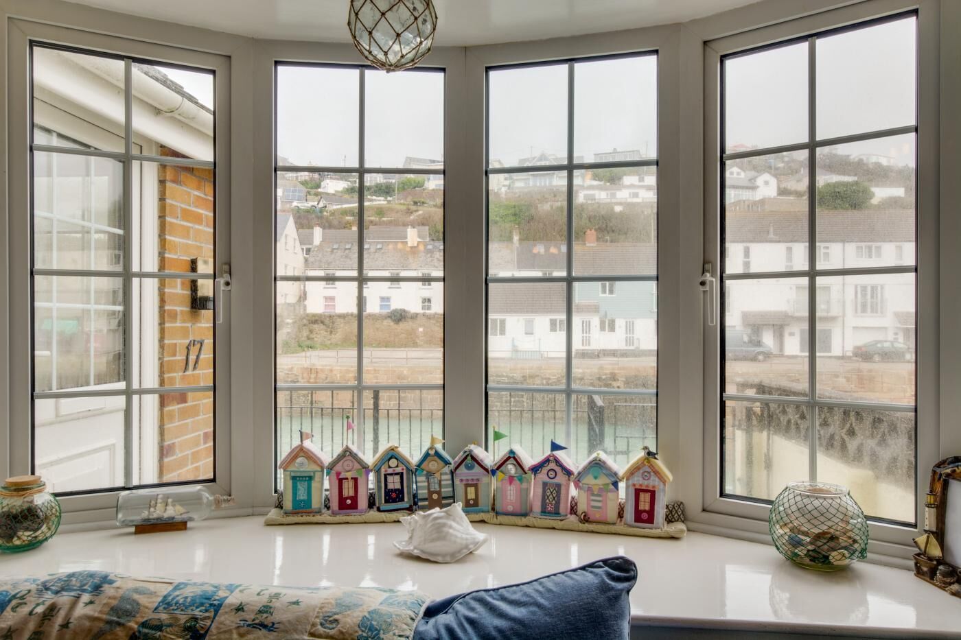 A room with a bay window overlooking a coastal area, featuring a row of colorful miniature houses on the windowsill.